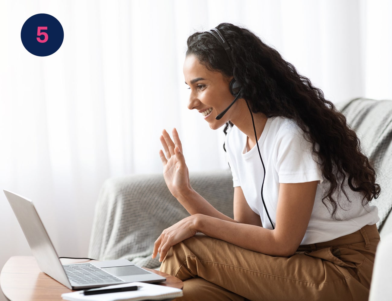 A remote worker sitting on a sofa and wearing a headset waves and smiles to her laptop screen
